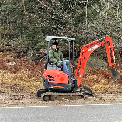 A man operates a red excavator parked on the roadside, engaged in construction work.